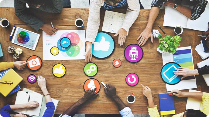 People sit around a wooden table working with colorful icons and charts, indicating collaboration and brainstorming in a team meeting.