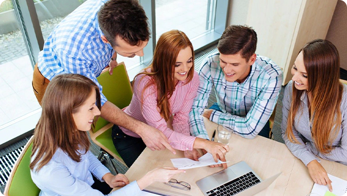 A group of five people sitting around a table, looking at documents and a laptop, engaged in discussion.